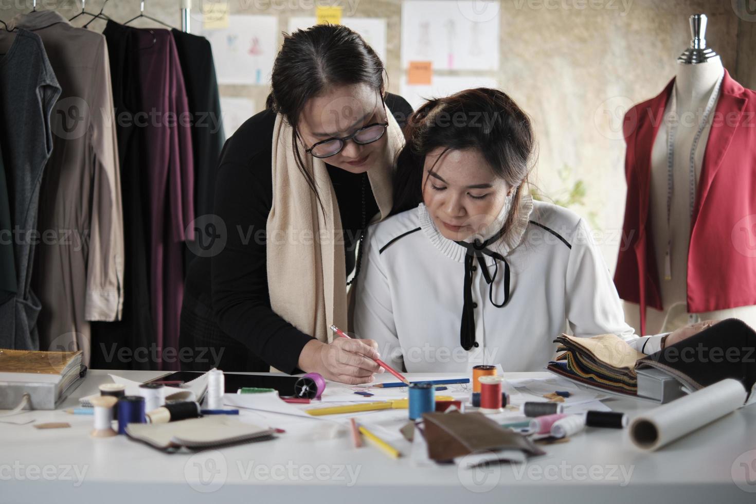 una diseñadora de moda asiática de mediana edad le enseña a un joven sastre adolescente en estudio con hilo colorido y tela de costura para ideas de colección de diseño de vestidos, pequeña empresa boutique profesional. foto