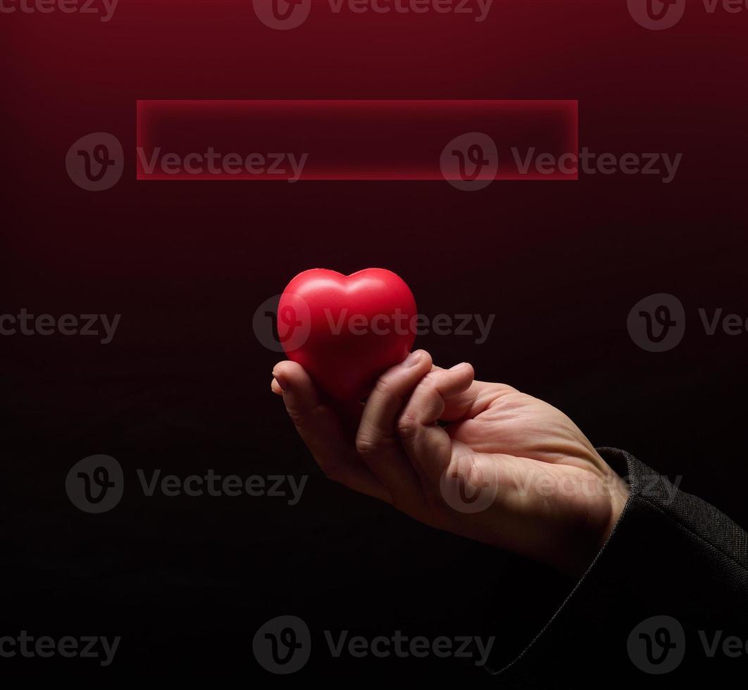 A man's hand holds a red heart on a dark background, a place for an inscription photo