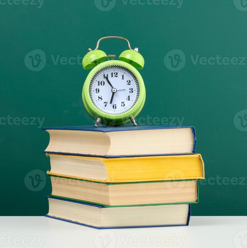 round alarm clock and stack of books on empty chalk green board background photo