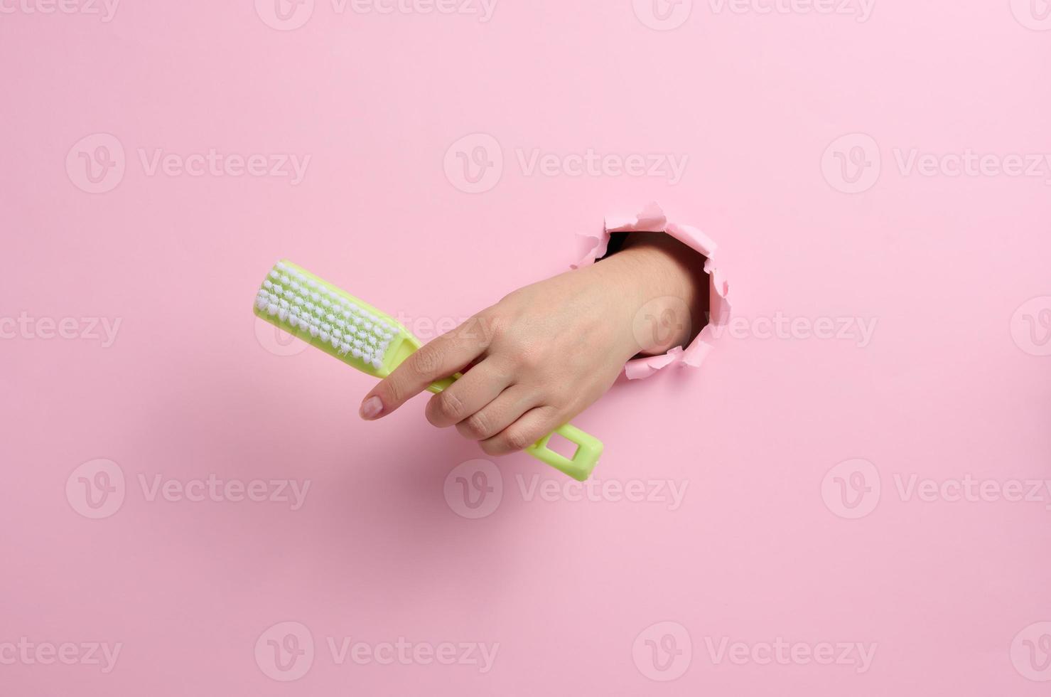 female hand holds a green plastic brush on a pink background. A part of the body sticks out of a hole with torn edges in a paper background photo