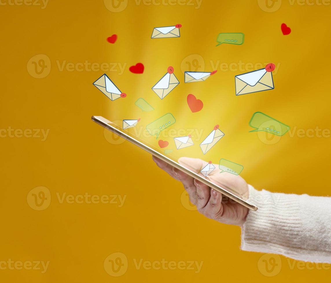 female hand holds a tablet with flying out hearts, dialogues and letters on a yellow background. Social media addiction, receiving correspondence and appraisal. Multitasking photo