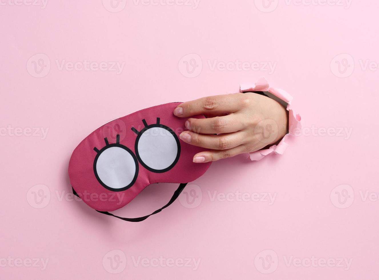 pink textile sleep mask in a female hand sticking out of a bull in a pink paper background, insomnia photo