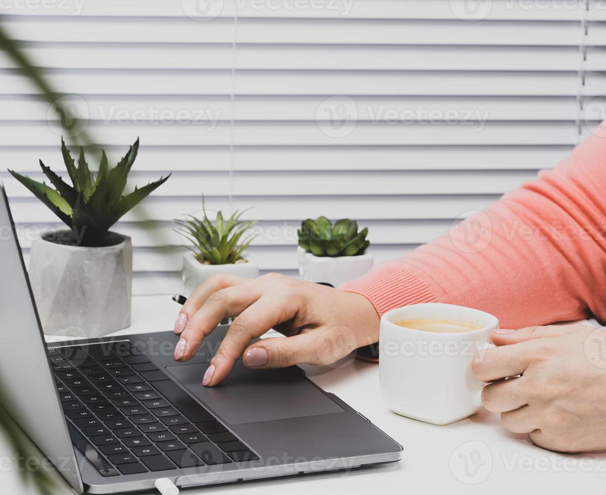woman in a pink sweater works on a laptop, in hand a white cup with coffee. Distance learning and work. Browse sites in a cafe photo