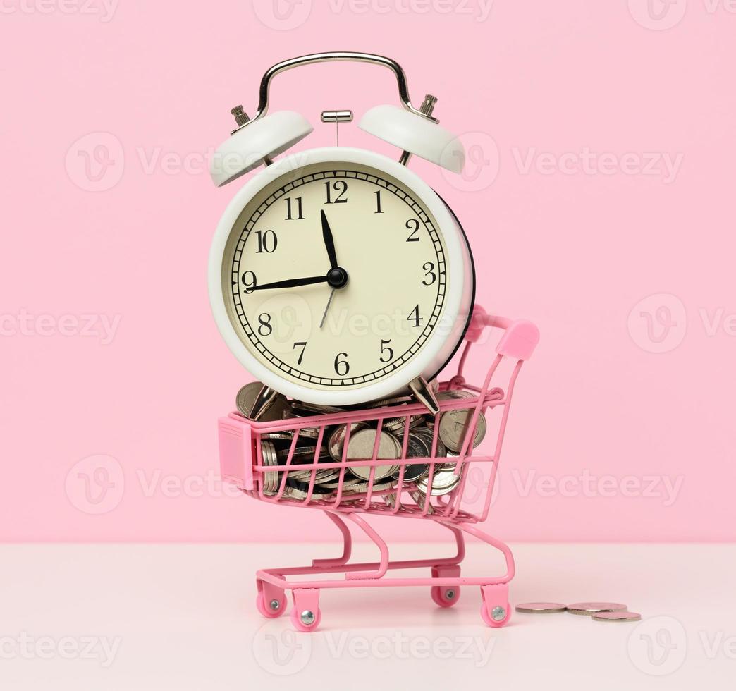 redondo alarma reloj en un miniatura compras carro con cambio en un blanco mesa. concepto hora es dinero, residuos de dinero y pobreza foto
