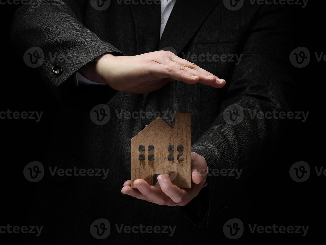 businessman in a suit holds a wooden model of a house on a dark background. Real estate sale and purchase concept, rent. Insurance and mortgage photo