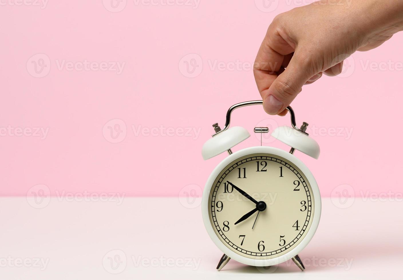 female hand holds a white metal alarm clock on a white table, time is ten to eight in the morning photo