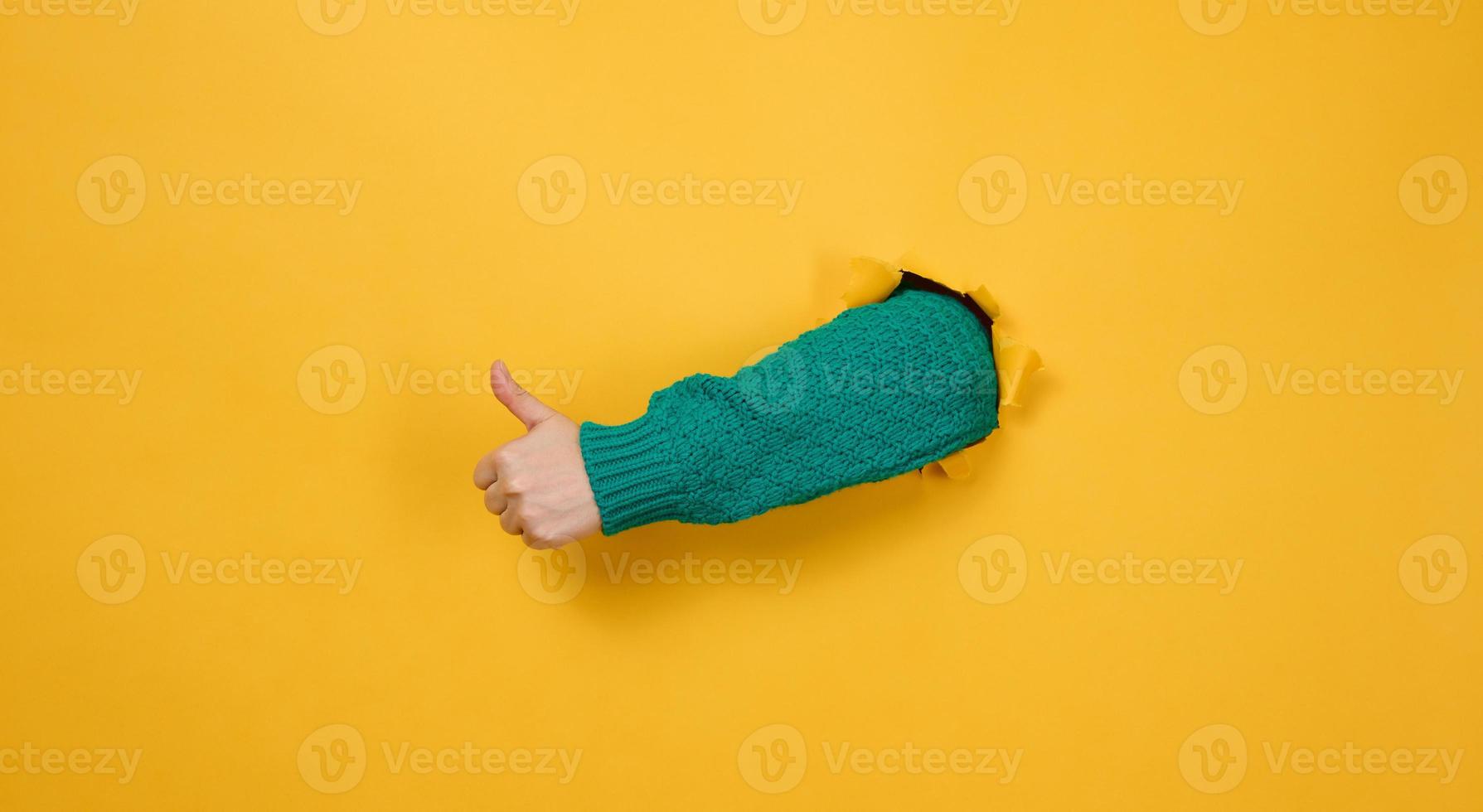 woman's hand sticks out of the torn hole and shows a gesture okay, thumb up. Yellow background photo