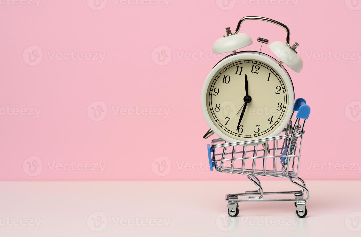 miniature metal shopping cart on wheels and in the middle of a round alarm clock on a white table. Start of discounts, sale photo