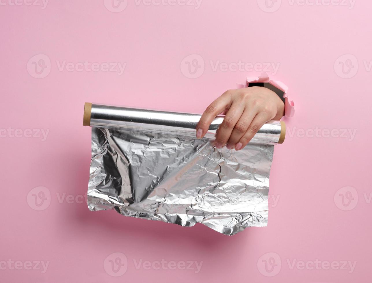 female hand holding a scroll of gray food foil on a pink background, part of the body sticking out of a torn hole photo