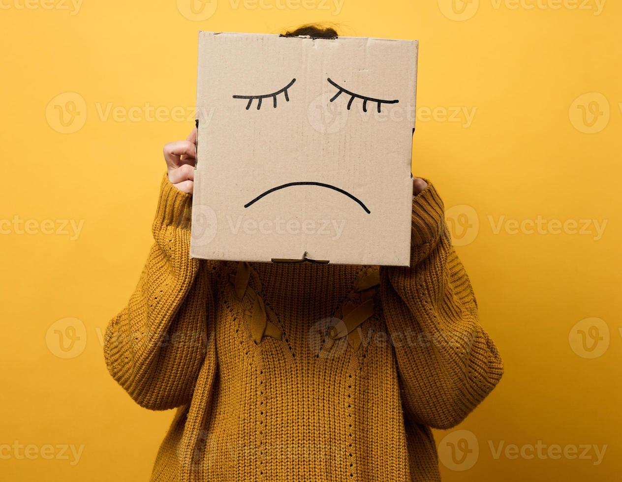 a woman in a brown sweater with a box on her head stands on a yellow background. Sad grimace, stress and depression concept. Mental health photo