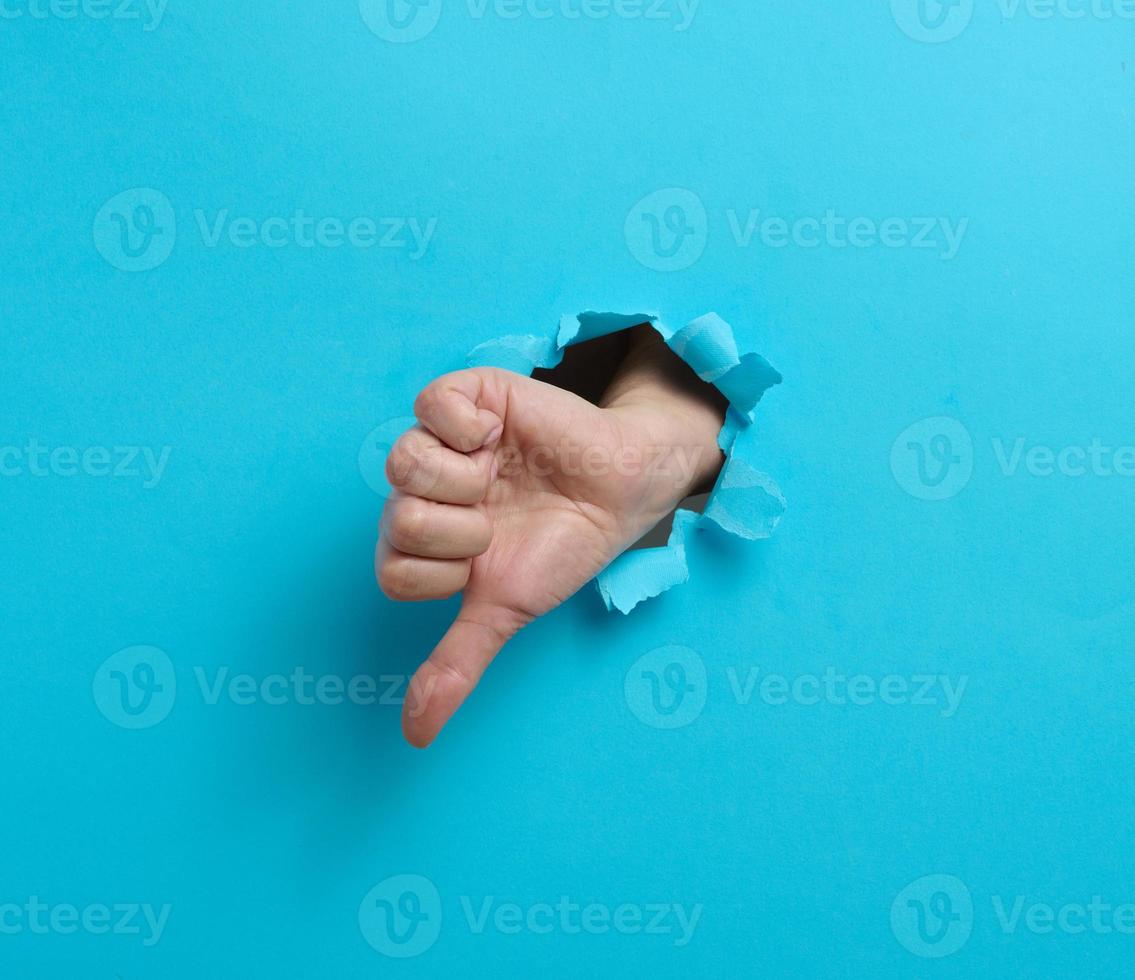 Dislike gesture break through the blue paper. A part of the body sticks out of a hole with ragged edges photo