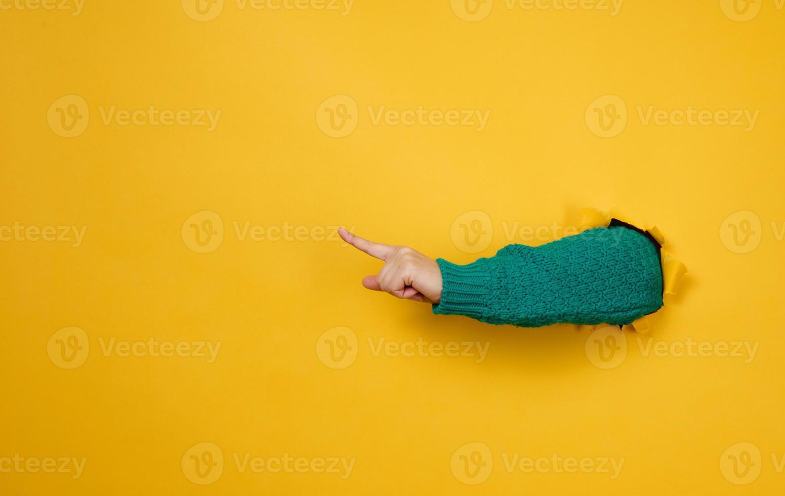 female hand sticks out of a torn hole in a yellow paper background, part of the body points with the index finger to the side. Copy space photo