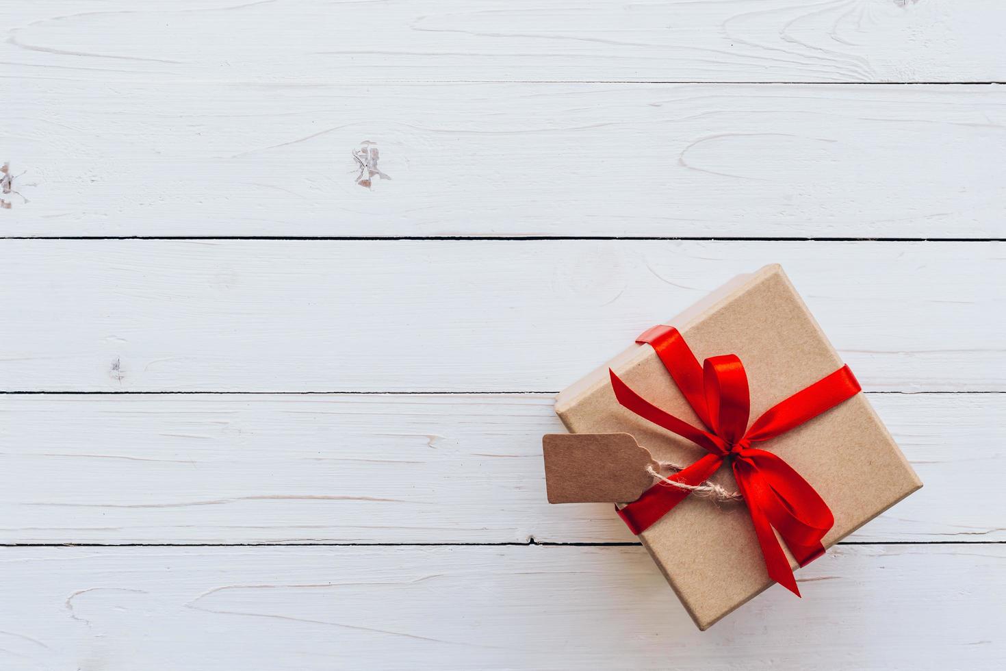 Above brown gift box with tag on wooden board background. Gift box with red ribbon on wooden white background with space. photo