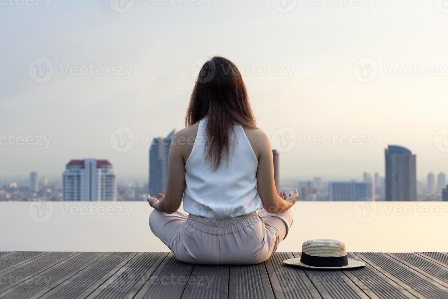Back of woman relaxingly practicing meditation at the swimming pool rooftop with the view of urban skyline building to attain happiness from inner peace wisdom concept photo