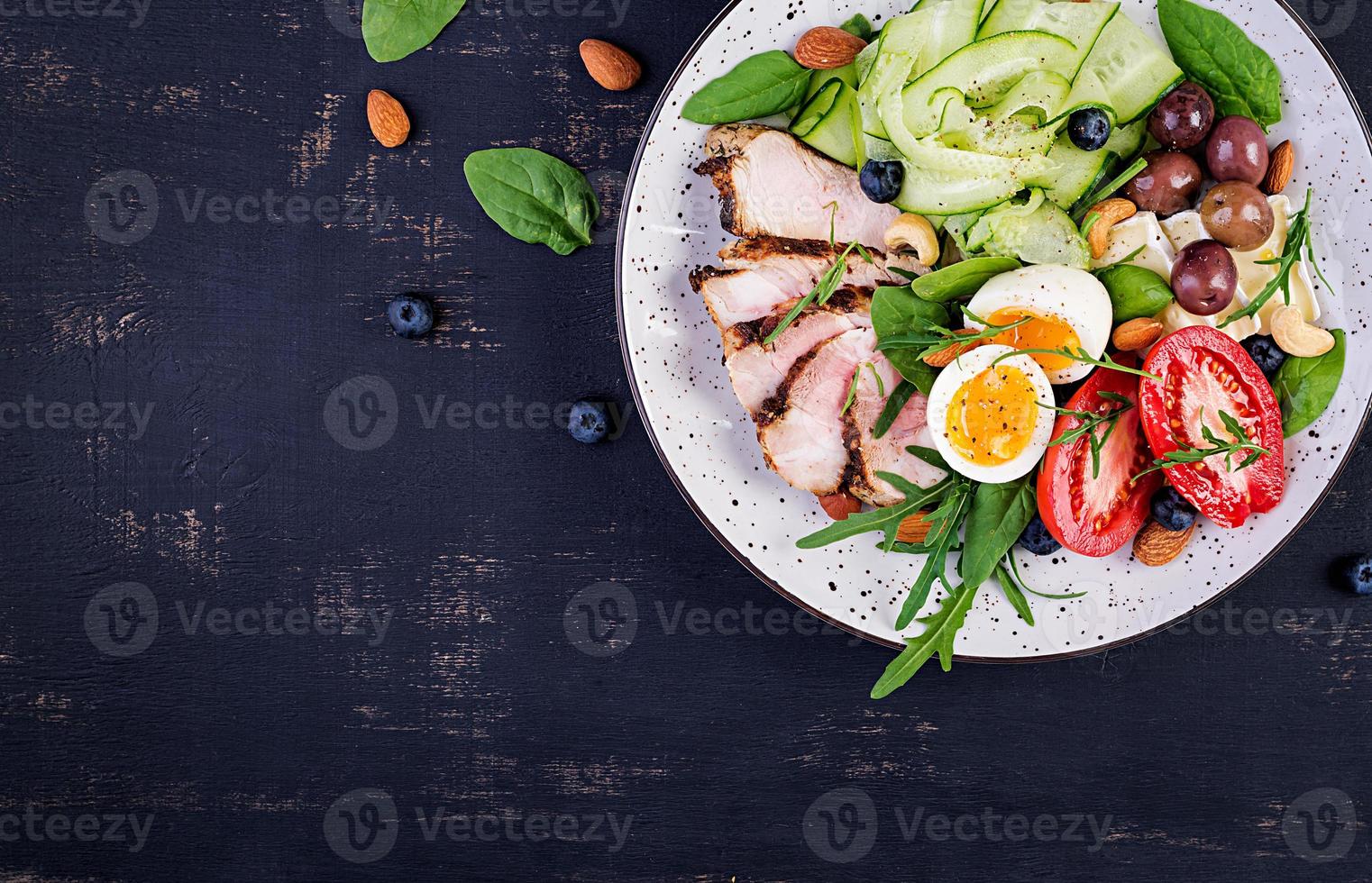 Ketogenic diet. Keto brunch. Boiled egg, pork steak and olives, cucumber, spinach, brie cheese, nuts and tomato. Top view photo