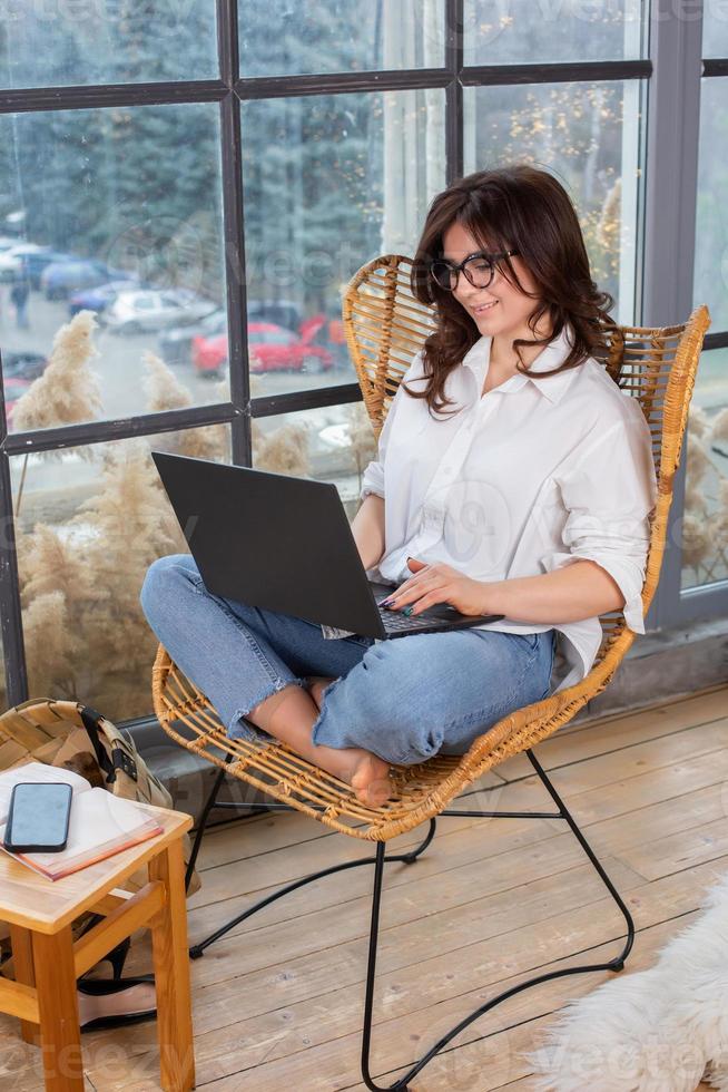 Beautiful businesswoman working on a laptop in a chair near the window. Female freelancer connecting to internet via computer. Blogger or journalist writing new article. photo
