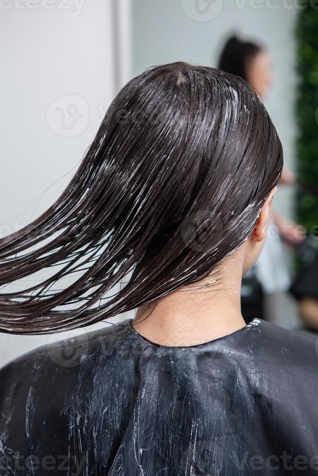 Hairdresser applies a hair mask to straight black hair. Hair care at the beauty salon. photo