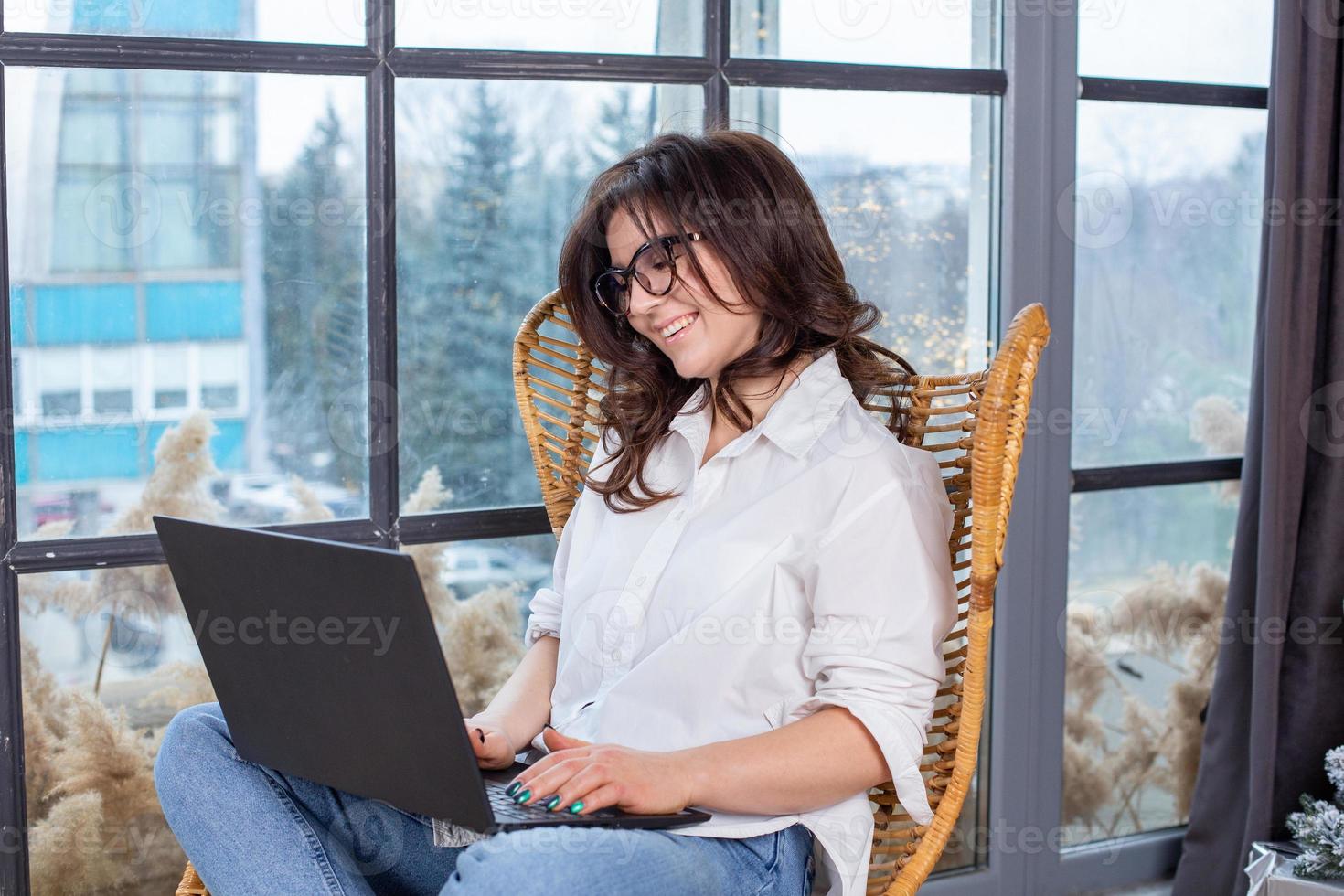 Beautiful businesswoman working on a laptop in a chair near the window. Female freelancer connecting to internet via computer. Blogger or journalist writing new article. photo