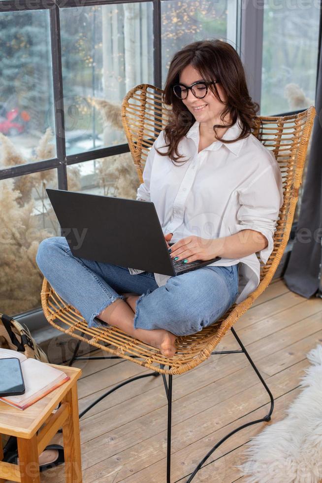 Beautiful businesswoman working on a laptop in a chair near the window. Female freelancer connecting to internet via computer. Blogger or journalist writing new article. photo