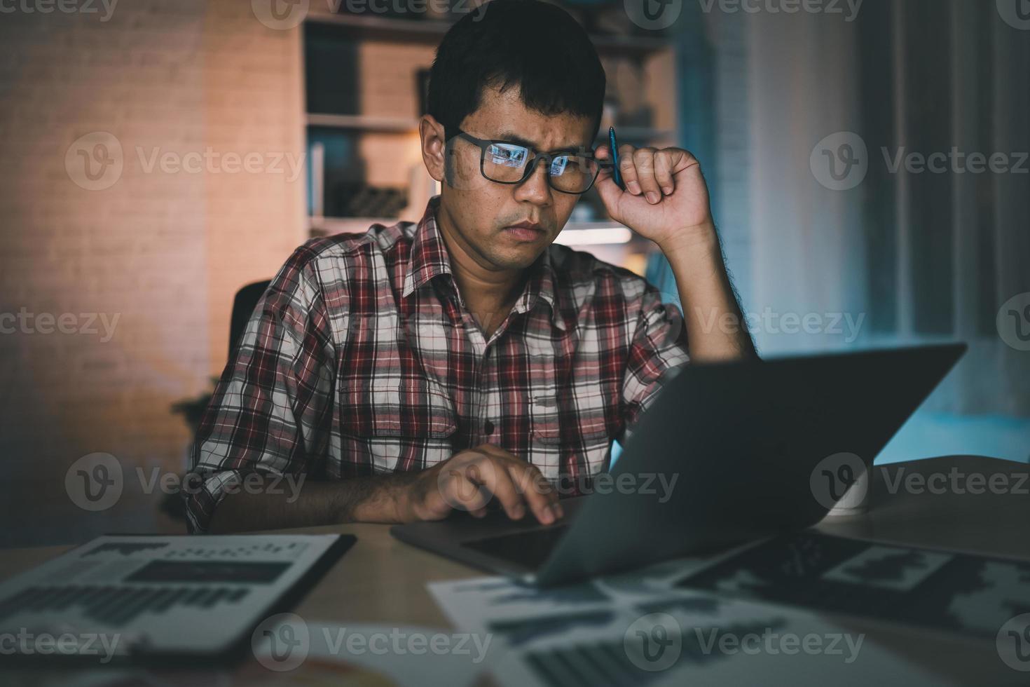 Asian freelance man having stressful depression sad time working on laptop home night. Depression man sad serios working from home. Man wearing plaid shirt red is rushing work overtime for deadline. photo