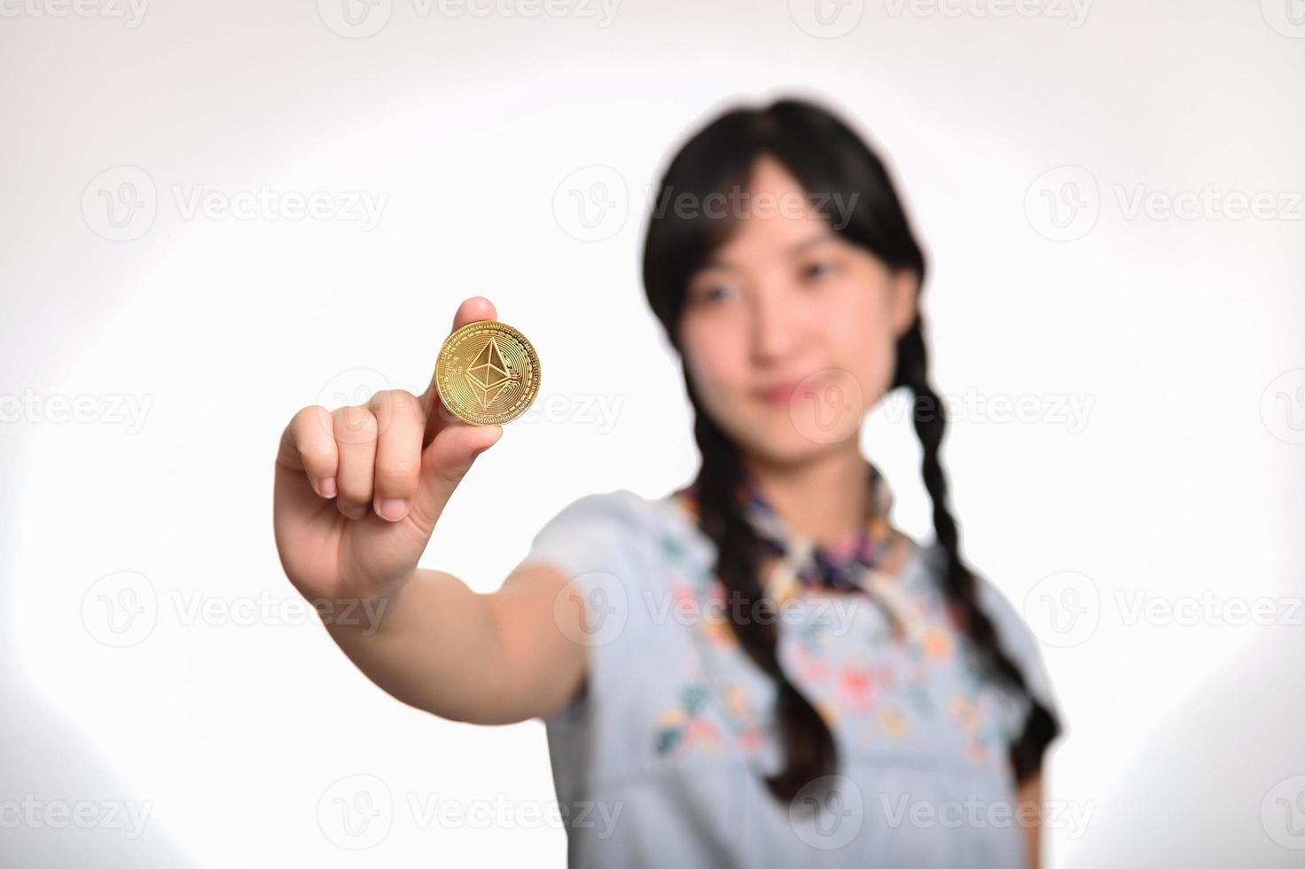 Portrait of beautiful happy young asian woman in denim dress holding crypto currency coin on white background. photo