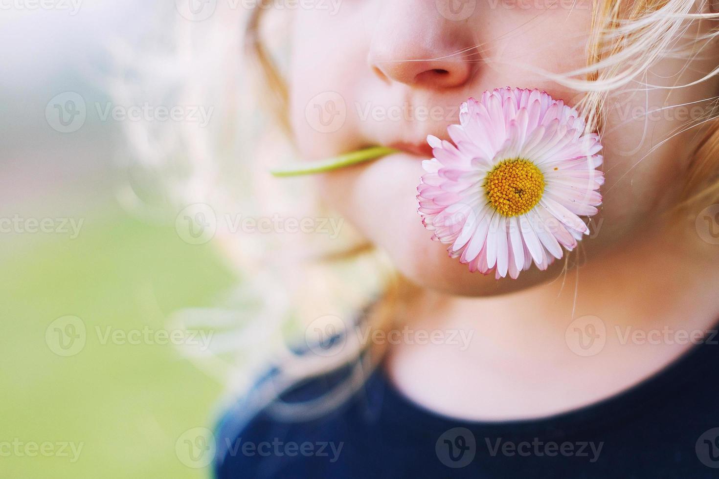 niña con un margarita flor en su boca en un verde antecedentes cerca. labios con flor en césped antecedentes. retrato de un pequeño niña foto