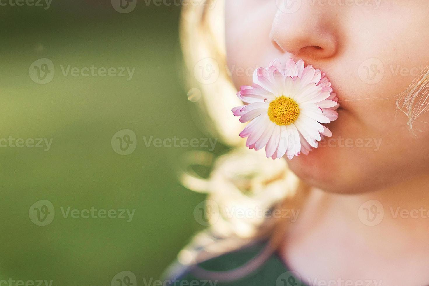 niña con un margarita flor en su boca en un verde antecedentes cerca. labios con flor en césped antecedentes. retrato de un pequeño niña foto