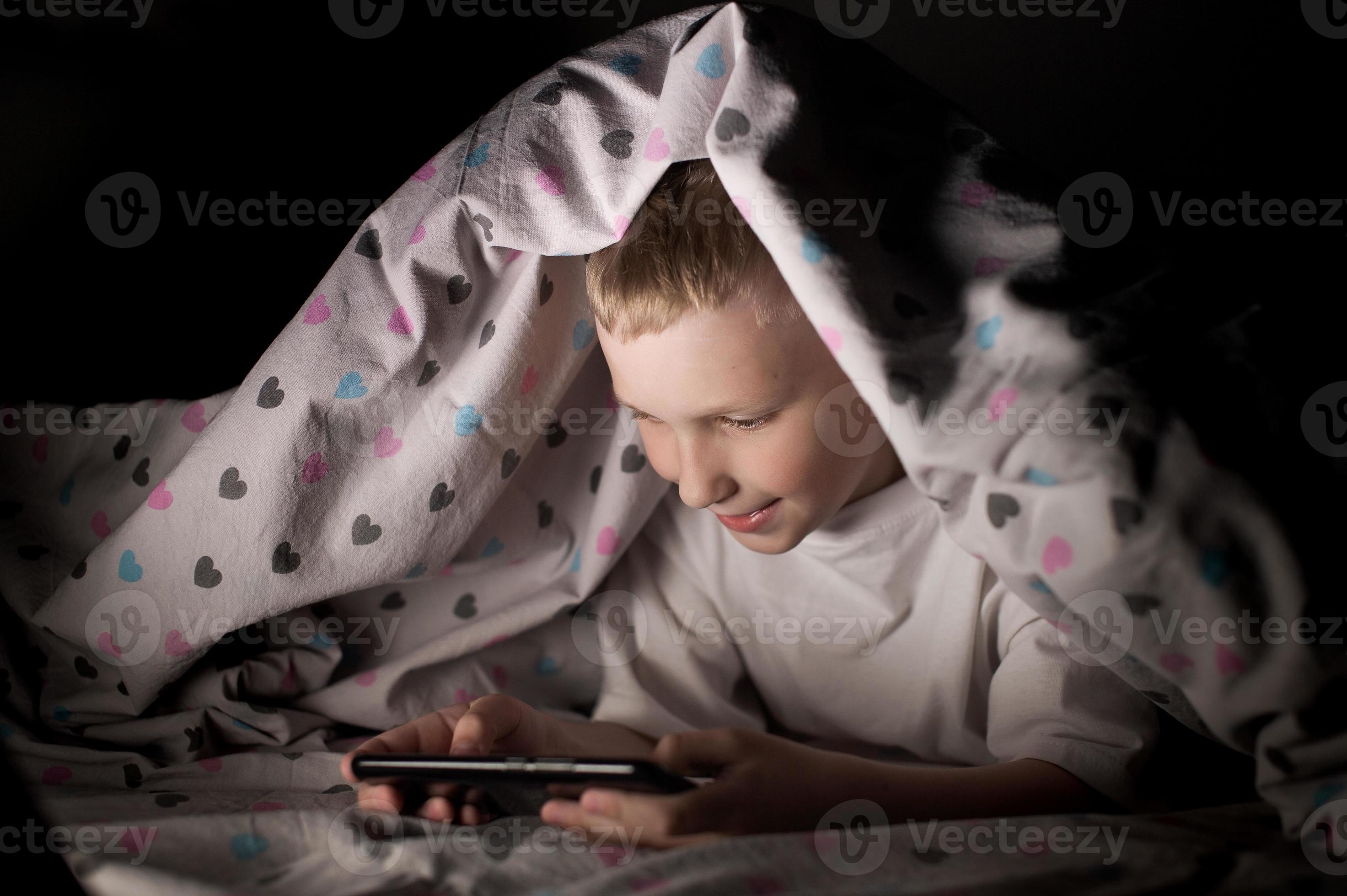 A little boy lies under a blanket at night playing on the phone