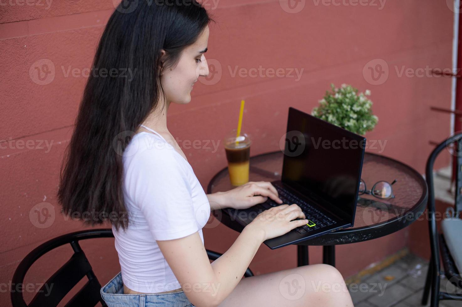 A young woman is sitting on the terrace with a laptop and a phone photo