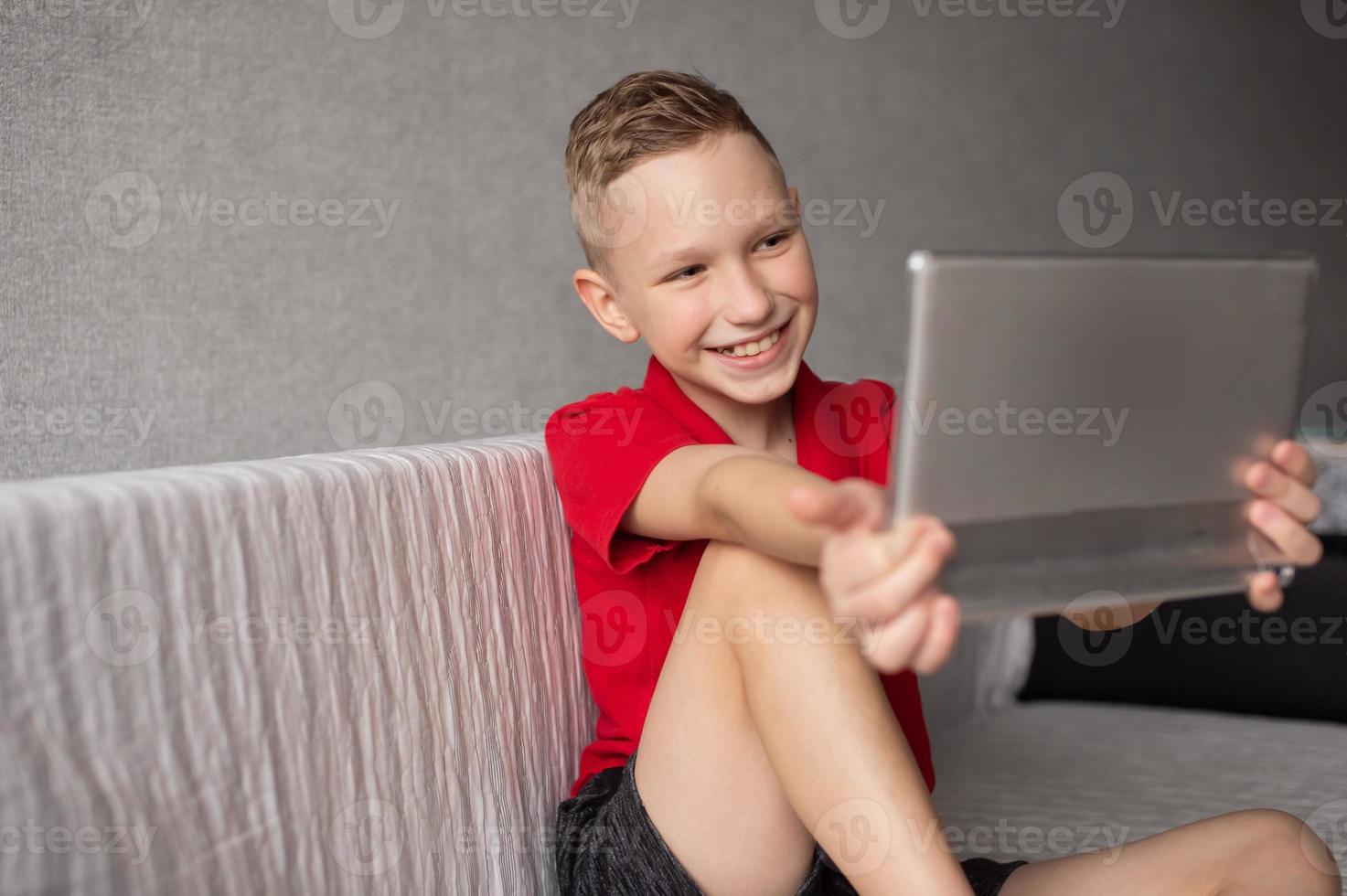 A happy boy in a red T-shirt is sitting on the couch with a tablet and laughing photo