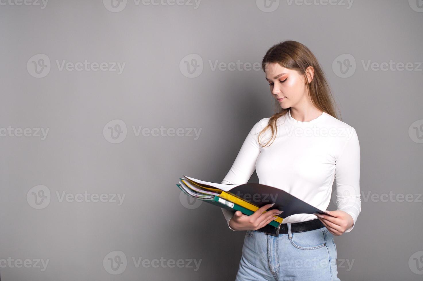 A cute student girl looks into an open folder for documents and stands on a gray background photo