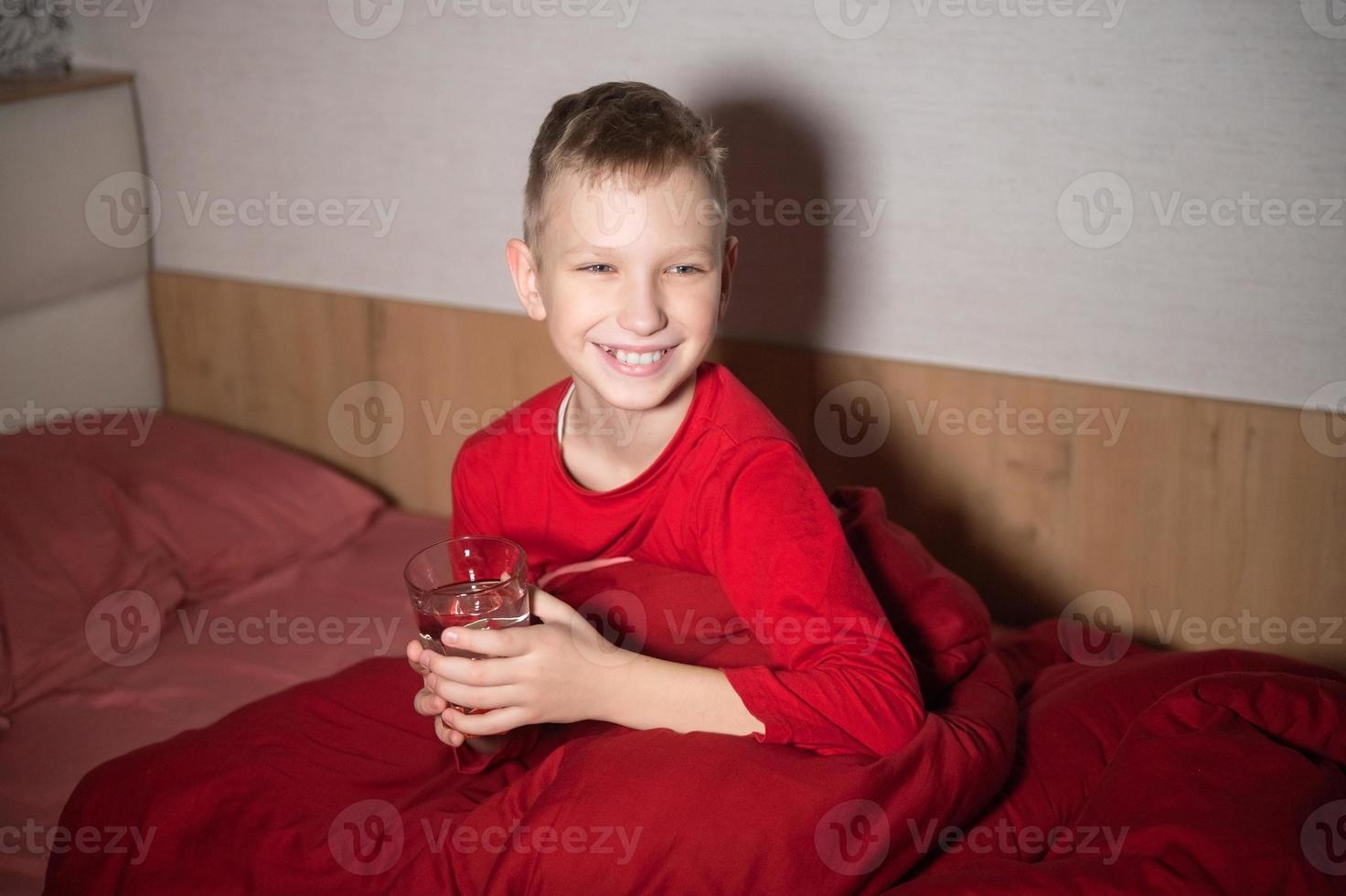 un contento chico es sentado en un rojo cama debajo un cobija con un vaso de agua foto