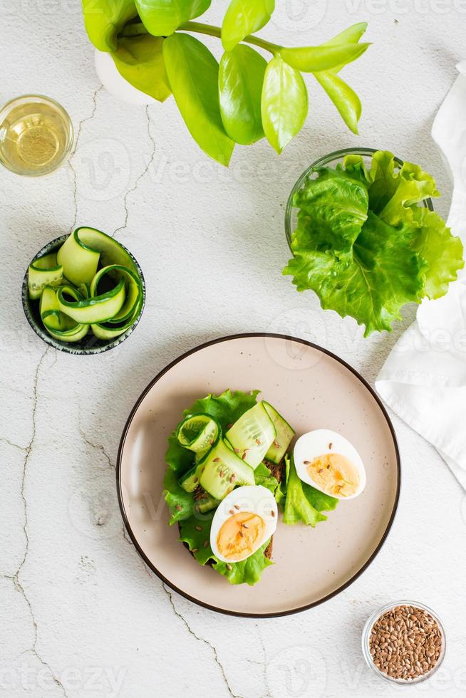 Ready to eat sandwich with rye bread, lettuce, cucumber, boiled egg and flax seeds on a plate on the table. Healthy eating on a diet. Top and vertical view. photo