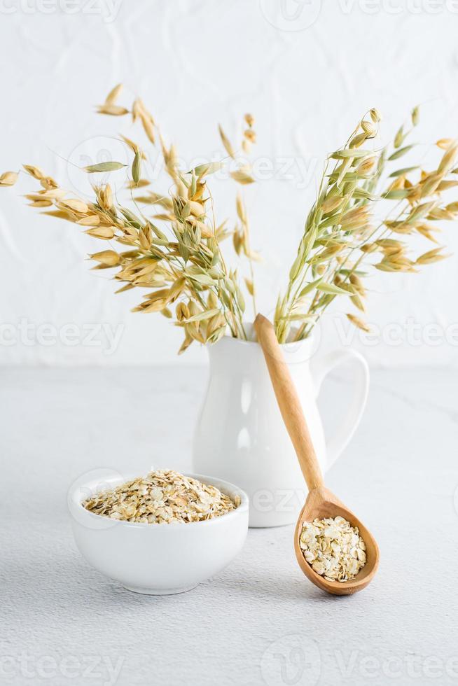 Dry oatmeal in a bowl and in a wooden spoon and a jug with ears on a light table. Homemade healthy diet. Vertical view photo