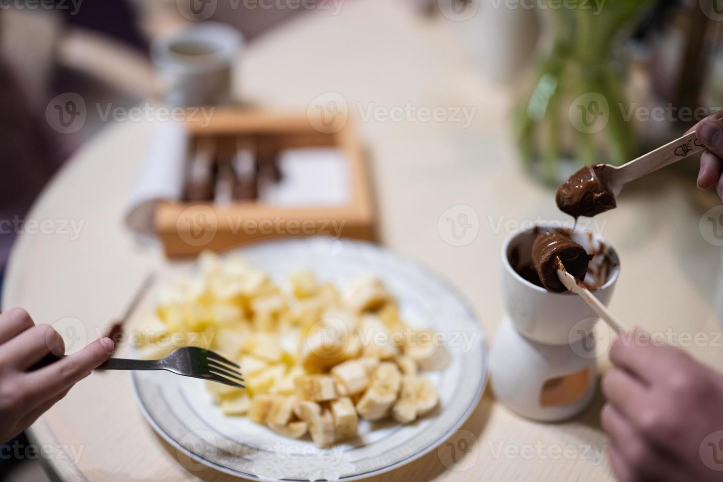Children eat fruits and desserts, drink tea at home in the evening kitchen. Close up hand with chocolate on a stick for melting. photo