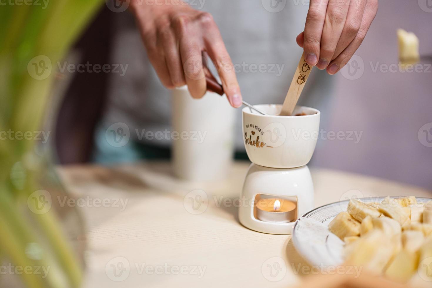 Children eat fruits and desserts, drink tea at home in the evening kitchen. Close up hand with chocolate on a stick for melting. photo