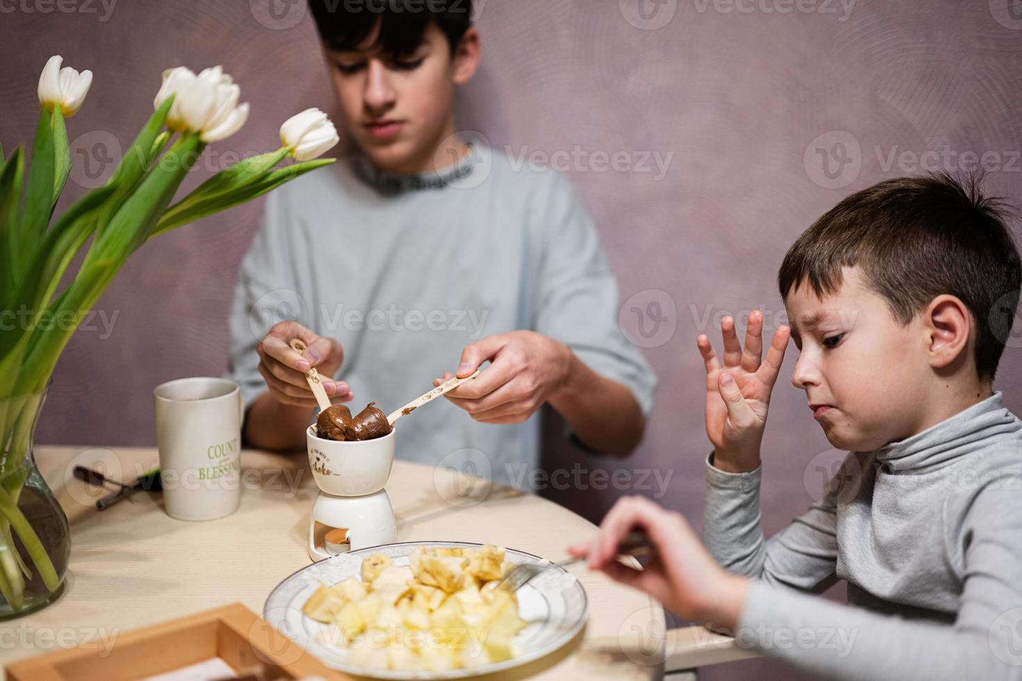 niños comer frutas y postres, bebida té a hogar en el noche cocina. chocolate en un palo para derritiendo. foto