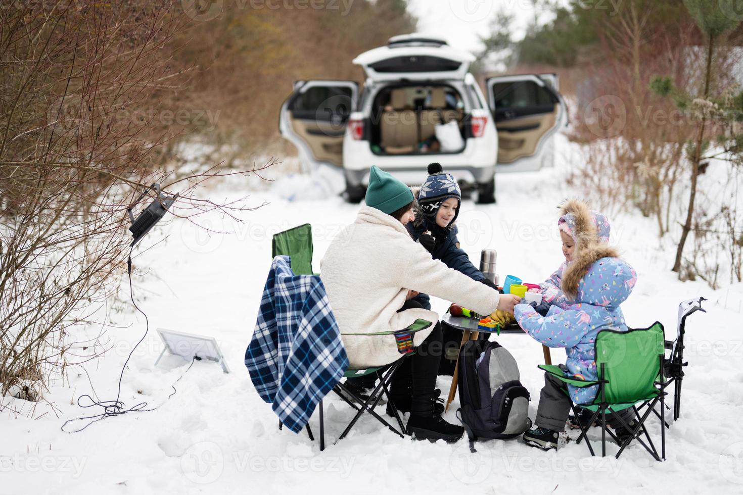 Young woman with children in winter forest on a picnic. Mother and three kids against they car with open trunk. photo