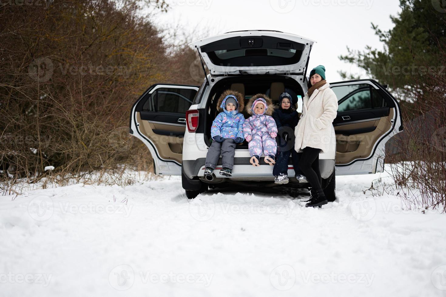 Mother with three children sit on car suv with open trunk stand in winter forest. photo