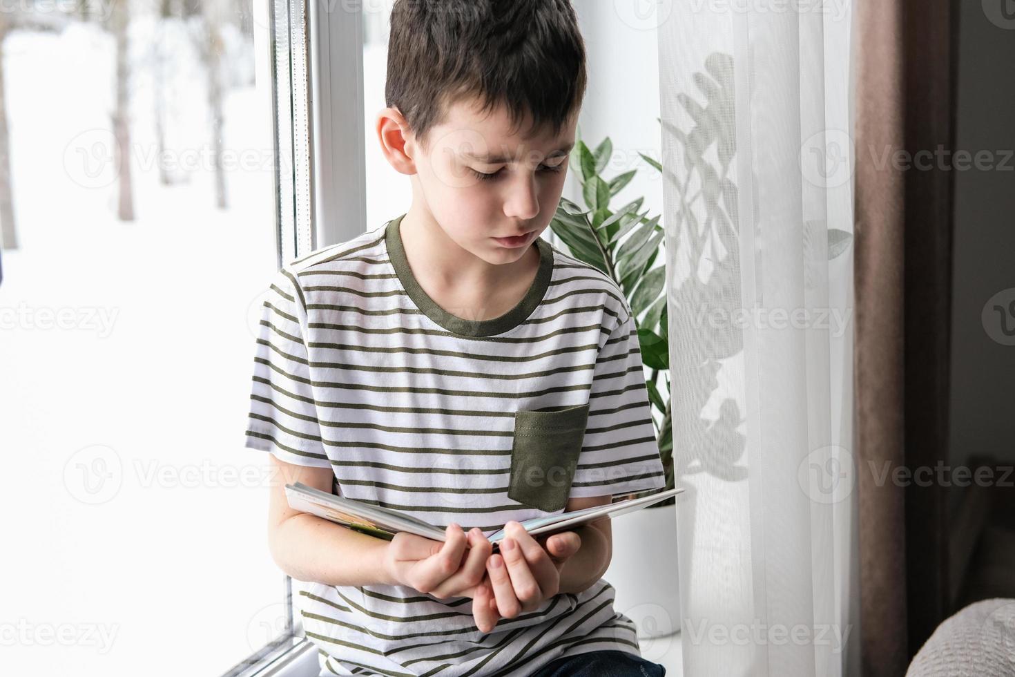 A six-year-old boy is reading a book while sitting by the window. Home schooling photo
