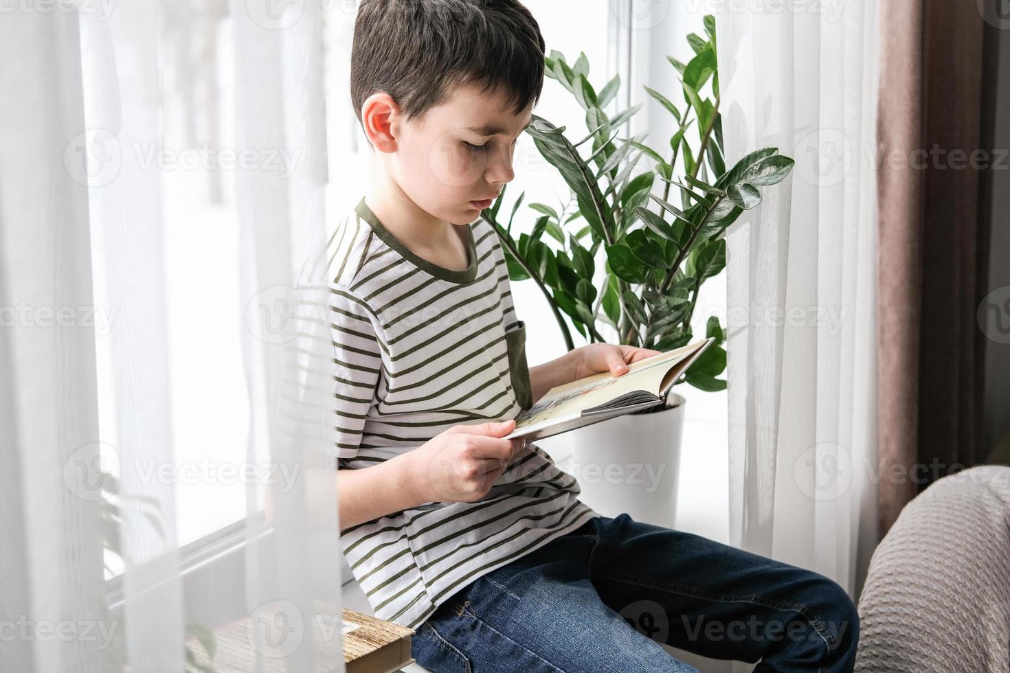 A six-year-old boy is reading a book while sitting by the window. Home schooling photo