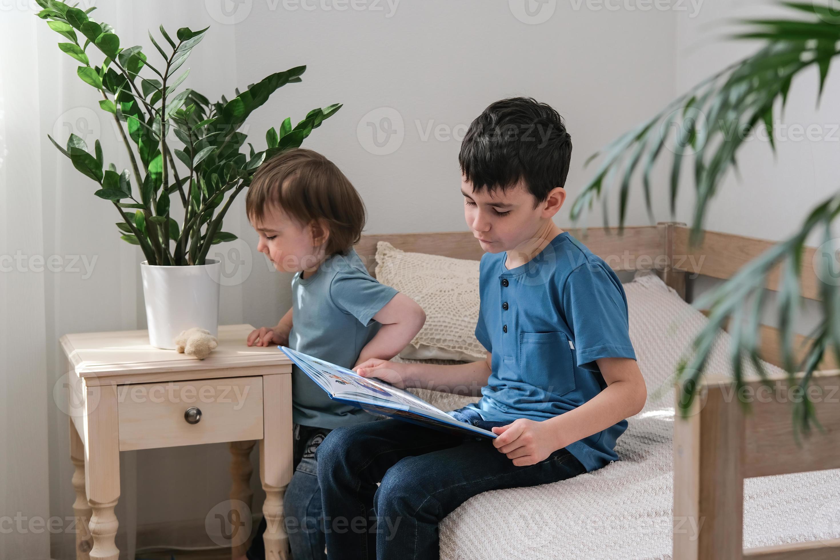 A sixyearold boy is reading a book while sitting on the bed. Home
