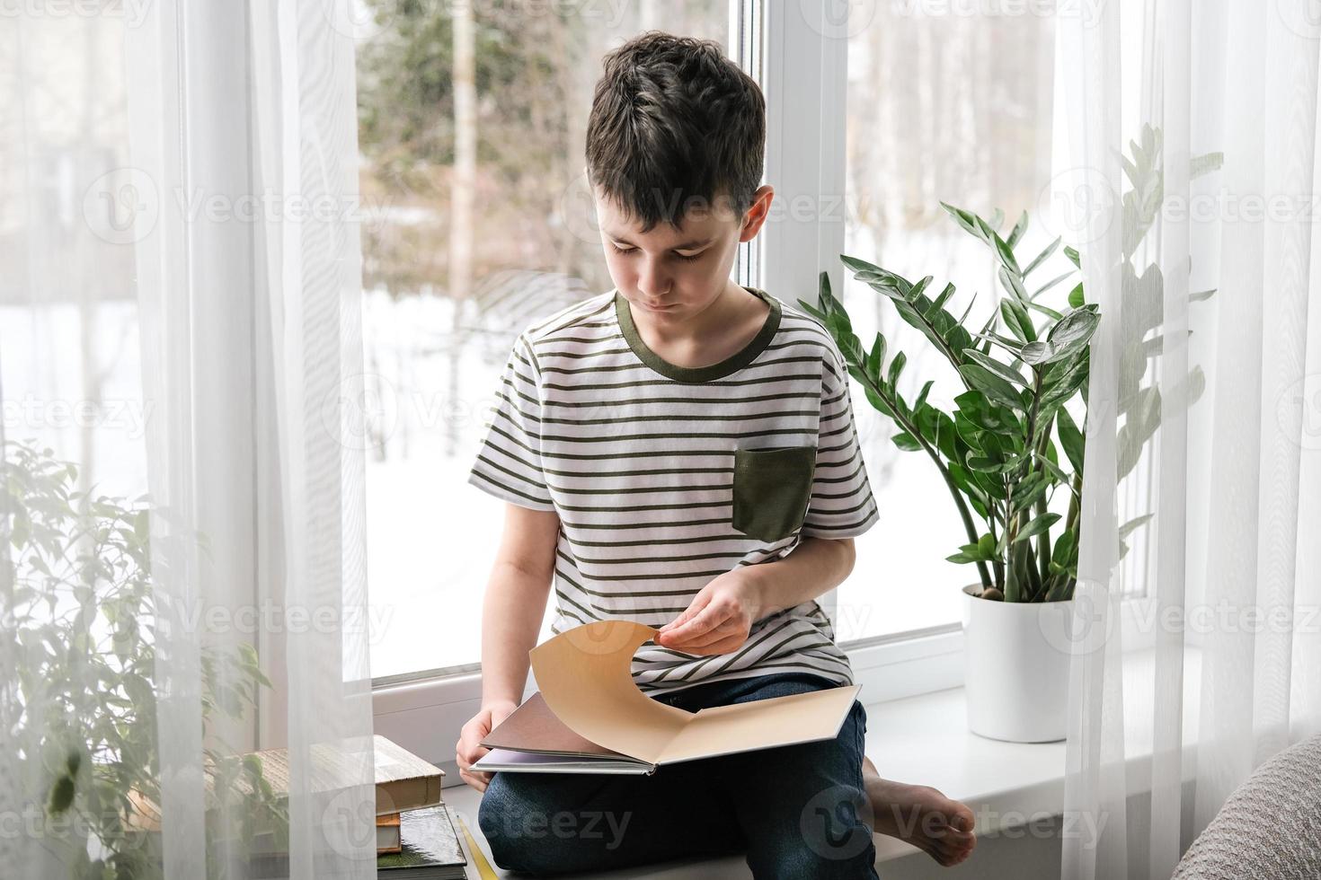 A six-year-old boy is reading a book while sitting by the window. Home schooling photo