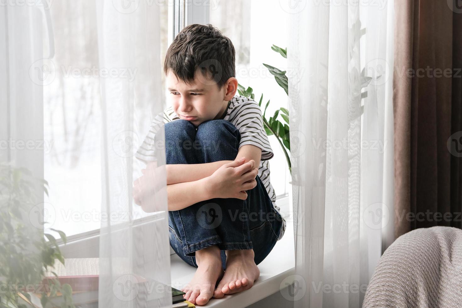 A sad boy sits on the windowsill hugging his knees. Bad mood, depression. The boy is sad alone at home photo