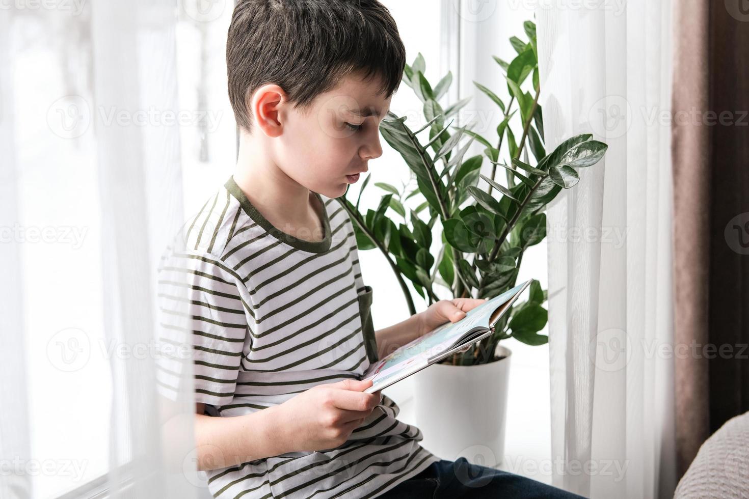 A six-year-old boy is reading a book while sitting by the window. Home schooling photo