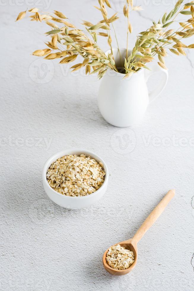 Dry oatmeal in a bowl and in a wooden spoon and a jug with ears on a light table. Home healthy food.  Vertical view photo