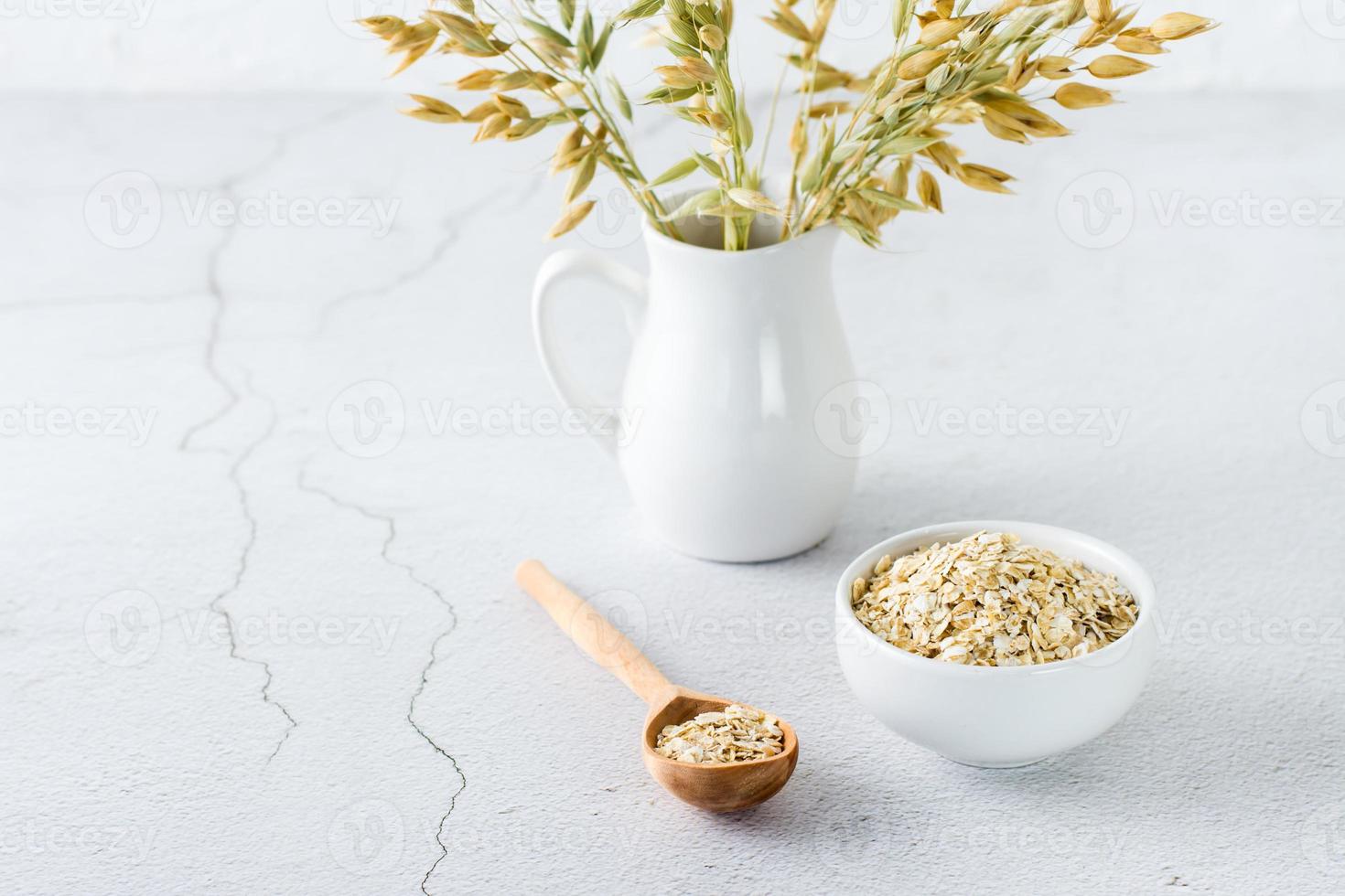 Dry oatmeal in a bowl and in a wooden spoon and a jug with ears on a light table. Home healthy food.  Copy space photo