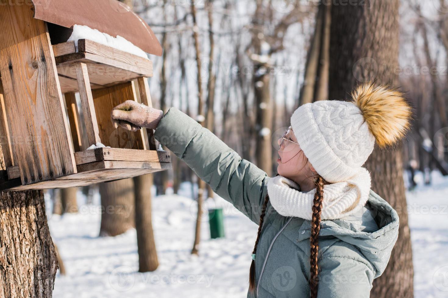 A teenager pours seeds into a homemade bird feeder on a tree in a winter park. Animal care. photo