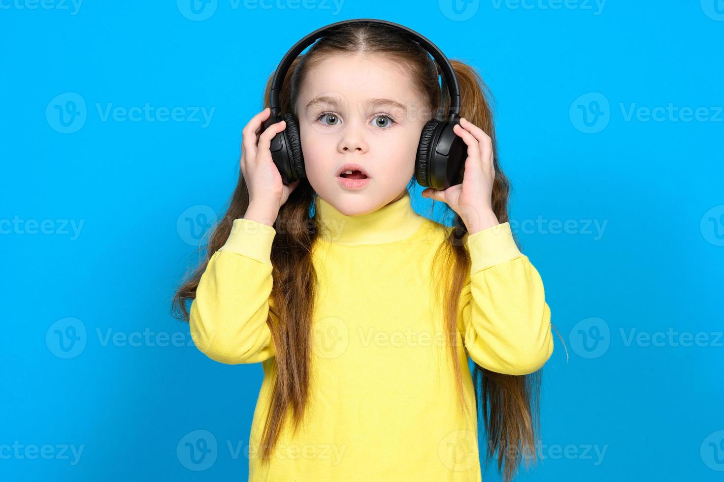 un pequeño niña en un amarillo suéter en un azul antecedentes escucha a música en inalámbrico auriculares foto