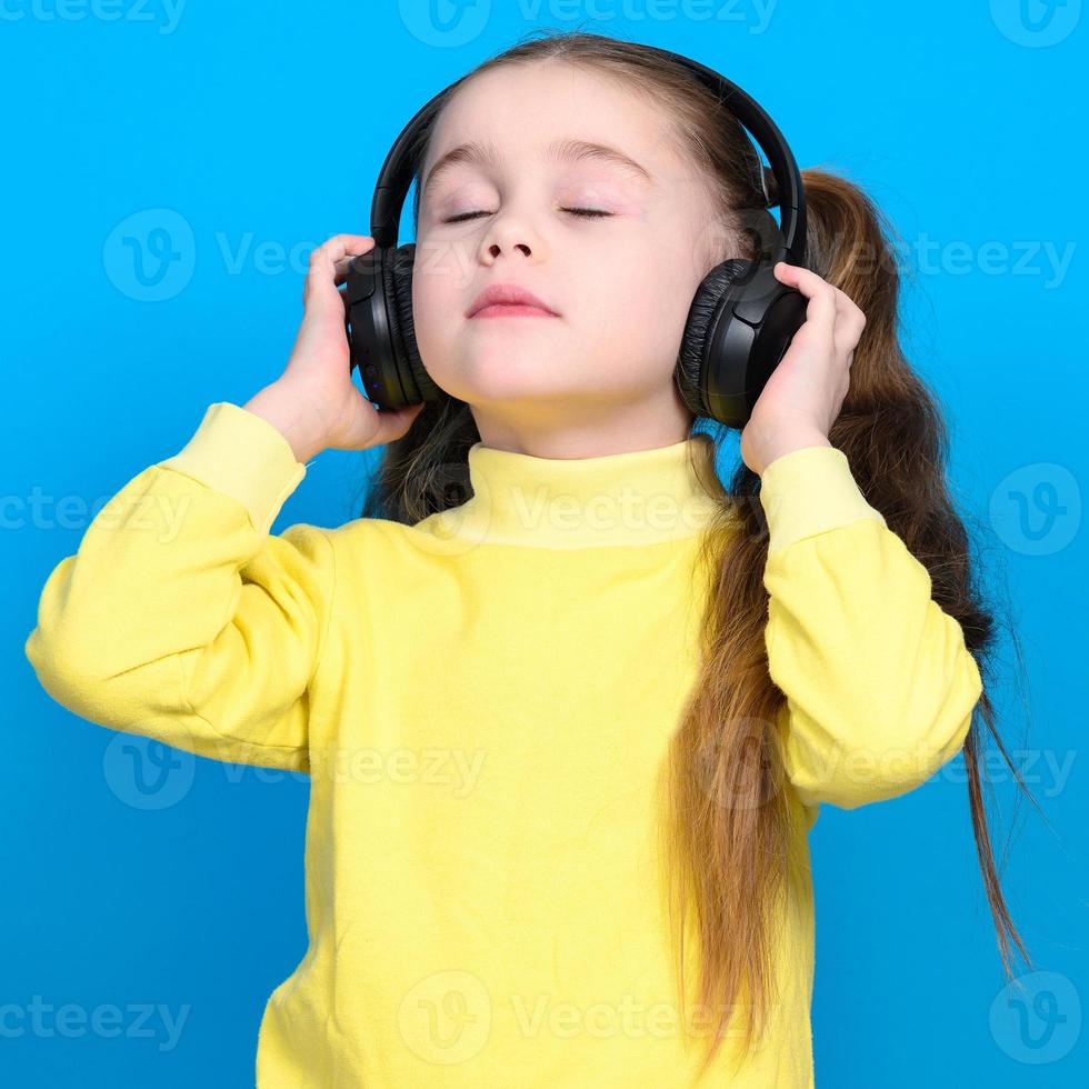 retrato de un niña en un azul antecedentes en un amarillo suéter, un pequeño niña con inalámbrico auriculares. foto